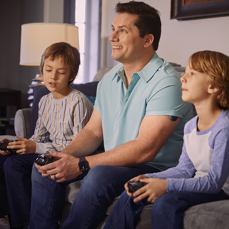 Hombre jugando a videojuegos con sus hijos mientras lleva un sistema de MCG implantable en el brazo.